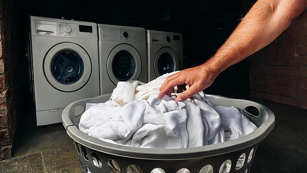 männerhand greift in einen großen gefüllten wäschekorb aus plastik, in dem sich weiße damenkleidung befindet, hintergrund: ein dunkler keller mit drei alten waschmaschinen
