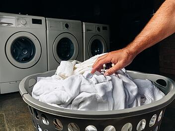 m&auml;nnerhand greift in einen gro&szlig;en gef&uuml;llten w&auml;schekorb aus plastik, in dem sich wei&szlig;e damenkleidung befindet, hintergrund: ein dunkler keller mit drei alten waschmaschinen
