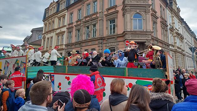 Ein Faschingsumzug mit verkleideten Musikern auf einem Wagen spielt vor Publikum in einer Stadt mit historischen Geb&auml;uden. Zuschauer tragen bunte Kost&uuml;me, und es herrscht fr&ouml;hliche Stimmung.