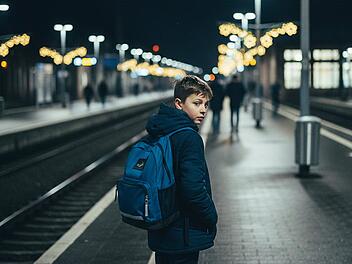 Erstelle realistisches Foto (16:9) einer Szene am Bahnhof Schwandorf in der Oberpfalz, Deutschland, aufgenommen an einem dunklen Winterabend. Im Vordergrund steht ein junger Junge mit Schulrucksack und Winterjacke, der mit gesenktem Blick und unsicherer K&ouml;rperhaltung am Rande des Bahnsteigs steht. Um ihn herum sind verschwommene Lichter der Weihnachtsdekoration und die k&uuml;hle Atmosph&auml;re eines typischen deutschen Bahnhofs im Dezember zu sp&uuml;ren. Die Aufnahme ist aus der Hand geschossen, mit starker K&ouml;rnung, sichtbarem Rauschen und leichtem Bewegungsunsch&auml;rfe, was dem Bild einen authentischen, ungeschliffenen Look verleiht. Im Hintergrund sind einige Passanten undeutlich zu erkennen, die ihrer Wege gehen, w&auml;hrend das Licht der Stra&szlig;enlaternen und die Reflexionen auf nassem Pflaster eine melancholische Stimmung erzeugen. Dokumentarischer Stil, 35-mm-Objektiv, leicht erh&ouml;hte Perspektive, spontane Alltagsszene in Deutschland, film grain, authentisch, winterliche Farbt&ouml;ne, keine Personen auf oder hinter der Theke sichtbar.