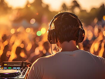 Ein DJ trägt Kopfhörer und steht an einem Mischpult. Im Hintergrund tanzt eine Menge bei Sonnenuntergang. Die Szene ist stimmungsvoll und sommerlich, passend zum "Vine & Vibes" in Bamberg.