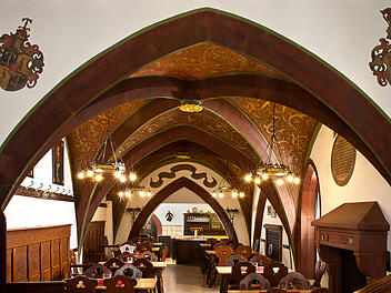 Ein traditionell eingerichtetes Gasthaus mit Gewölbedecken, Holztischen und -stühlen, geschmückt mit Wappen. Warmes, einladendes Licht sorgt für eine gemütliche Atmosphäre.