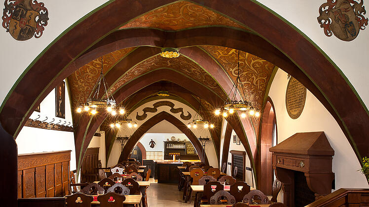 Ein traditionell eingerichtetes Gasthaus mit Gewölbedecken, Holztischen und -stühlen, geschmückt mit Wappen. Warmes, einladendes Licht sorgt für eine gemütliche Atmosphäre.