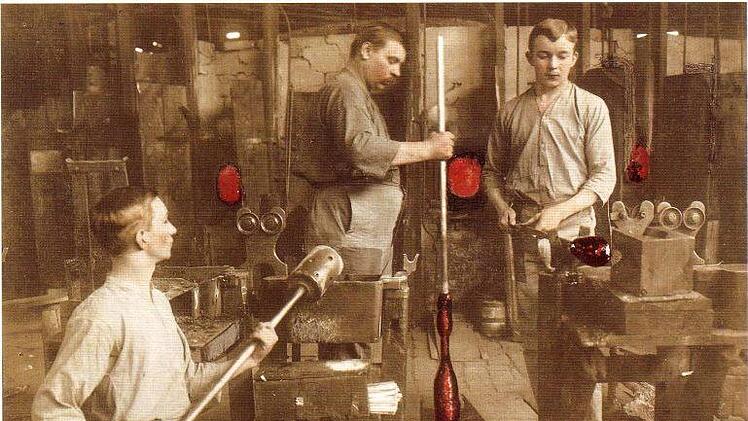 Hart und beschwerlich war die Arbeit der Glasbläser. Im Dreierteam arbeiteten sie an einer der 16 Werkstätten. Das Arbeitsergebnis: 300 Flaschen je Schicht. Die kolorierte Aufnahme entstand um 1925.  Repro: Gerd Fleischmann