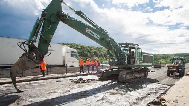 Ein Bagger schabt mit einem speziellen Schaufelaufsatz die Fahrbahndecke von der Brücke. Die Arbeiter und Ingenieure tragen Warnwesten. Foto: Hendrik Steffens