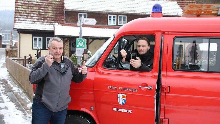 Mitten in Heiligkreuz gibt es zwar einen offiziellen Rettungstreffpunkt (Schild hinten), aber keinen Handy-Empfang: Kommandant Armin Henning (links) hat mit dem Digitalfunkgerät Empfang, während Daniel Niebusch (rechts), Vorsitzender der Wehr, mit dem Handy Pech hat. Foto: Ralf Ruppert