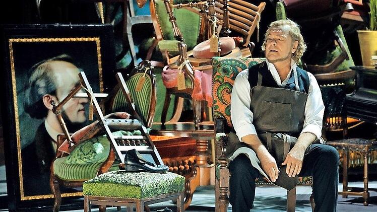 Der Bariton-Sänger Michael Volle als Hans Sachs in der Schusterstube des 2. Akts, hinter der sich das Wahnfried-Mobiliar des 1. Akts als Konkursmasse auftürmt.  Foto: Bayreuther Festspiele/Enrico Nawrath