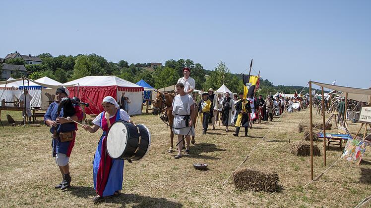 Menschen in mittelalterlichen Kostümen marschieren auf einem Fest mit Zelten und Fahnen. Eine Person führt eine Trommel, während andere in der Menge folgen, darunter auch ein Reiter.