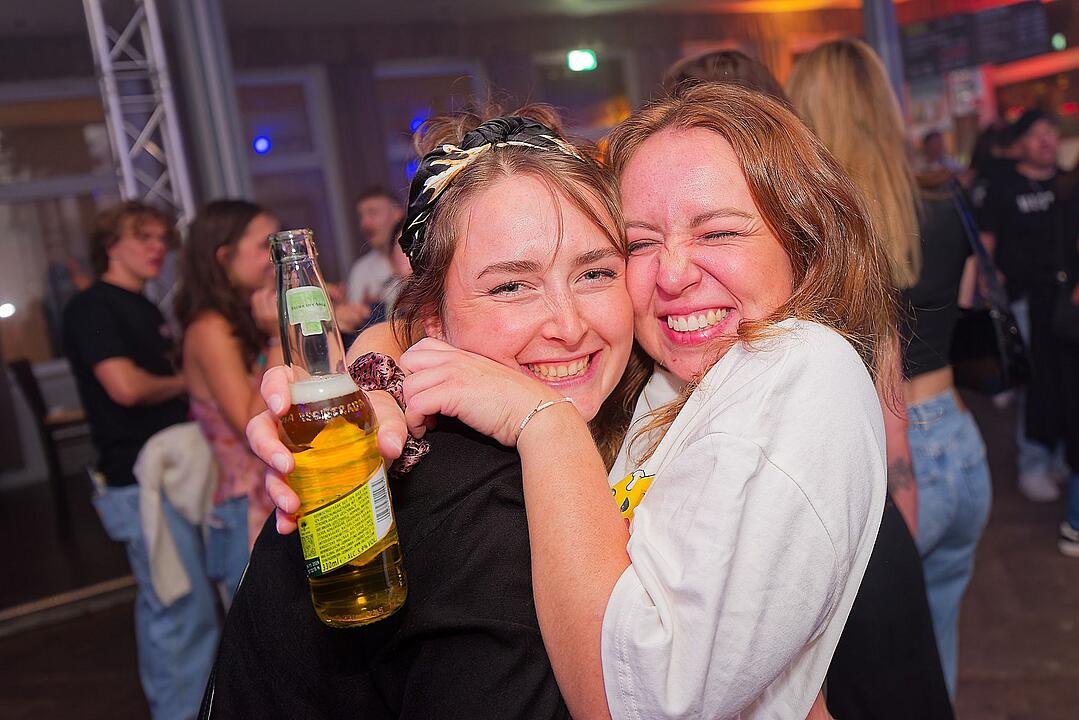Zwei fröhliche Personen umarmen sich auf einem Fest. Eine hält eine Flasche in der Hand. Im Hintergrund sind weitere feiernde Menschen zu sehen. Stimmung ist ausgelassen und lebhaft.