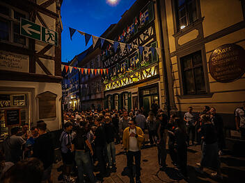 Eine belebte Gasse bei Nacht, dekoriert mit Wimpeln. Menschen stehen vor Fachwerkh&auml;usern und unterhalten sich. Stimmung eines lebhaften Festes, m&ouml;glicherweise die Bamberger Sandkirchweih.