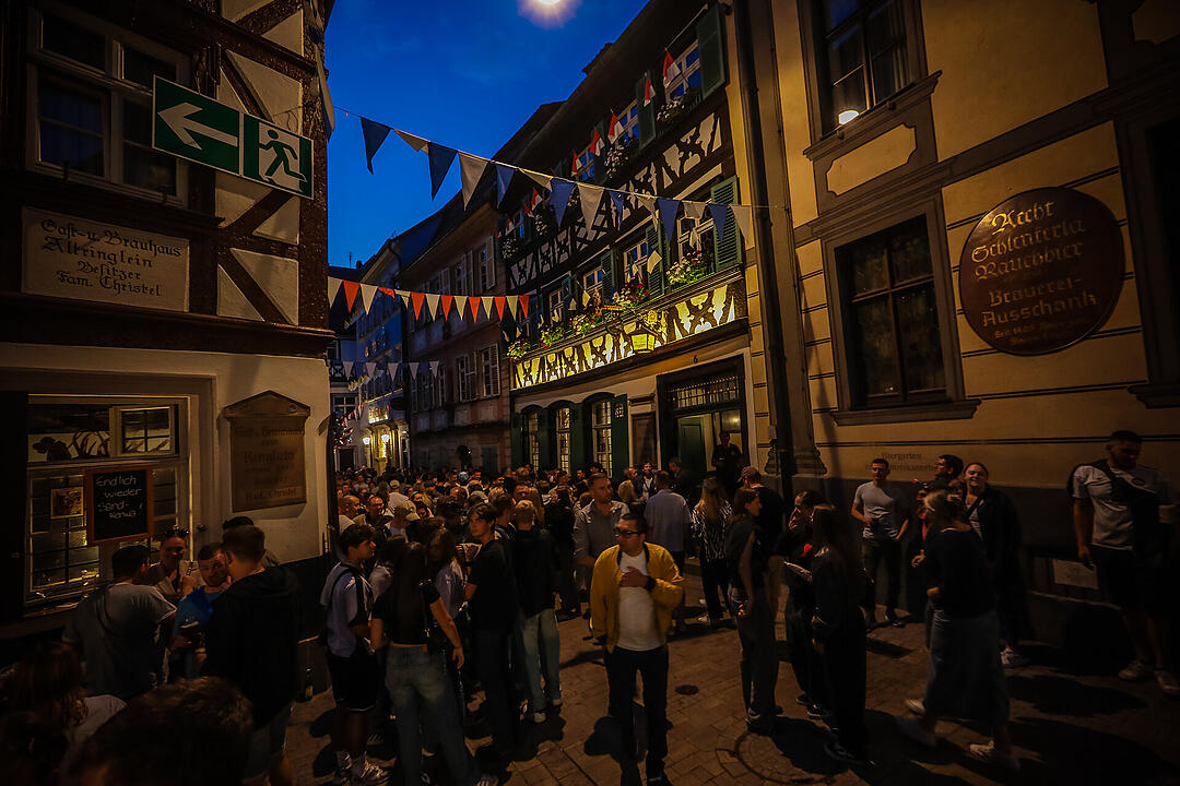 Eine belebte Gasse bei Nacht, dekoriert mit Wimpeln. Menschen stehen vor Fachwerkhäusern und unterhalten sich. Stimmung eines lebhaften Festes, möglicherweise die Bamberger Sandkirchweih.