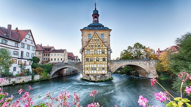 Fachwerkhaus auf Brücke über Fluss, umgeben von Altstadt und blühenden Blumen. Historischer Charme von Bamberg, UNESCO-Weltkulturerbe.