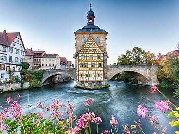 Fachwerkhaus auf Brücke über Fluss, umgeben von Altstadt und blühenden Blumen. Historischer Charme von Bamberg, UNESCO-Weltkulturerbe.