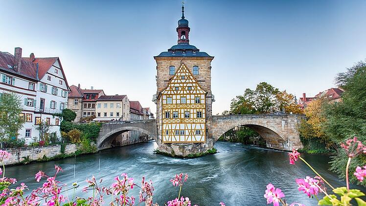 Fachwerkhaus auf Brücke über Fluss, umgeben von Altstadt und blühenden Blumen. Historischer Charme von Bamberg, UNESCO-Weltkulturerbe.
