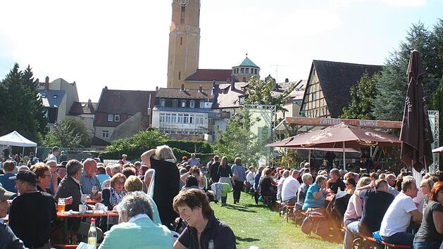 Eine Menschenmenge genießt ein Fest im Freien, sitzt an Biertischen. Im Hintergrund stehen Fachwerkhäuser und ein Kirchturm. Der Sonnenschein sorgt für eine fröhliche Atmosphäre.
