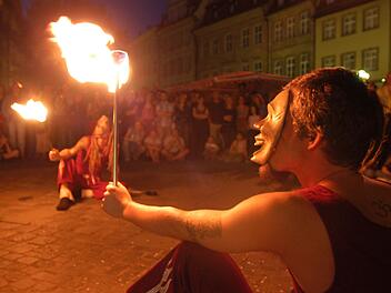 Ein Feuerspucker mit Maske hält eine brennende Fackel. Im Hintergrund sitzt ein weiterer Performer. Zuschauer umringen die Szene in einer stimmungsvoll beleuchteten Altstadt.