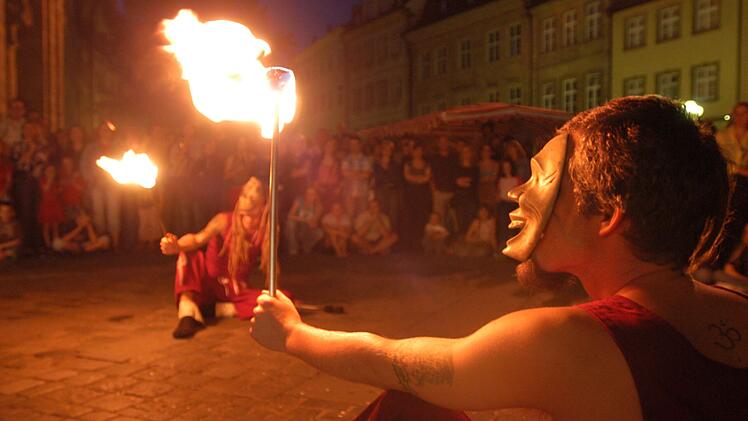 Ein Feuerspucker mit Maske hält eine brennende Fackel. Im Hintergrund sitzt ein weiterer Performer. Zuschauer umringen die Szene in einer stimmungsvoll beleuchteten Altstadt.