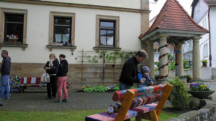 "Was ist denn mit der Bank passiert?" scheint sich der kleine Tim zu fragen. Der Dorfplatz in Gemünda präsentiert sich seit Sonntag in neuem Gewand. Auch der Brunnen hat ein Strickkleid bekommen. Foto: Bettina Knauth