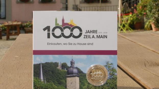 Die Jubiläumsmünze ist eingebettet in eine Klappkarte mit Zeiler Motiven und natürlich im Design der 1000-Jahr-Feier gestaltet.  Sabine Weinbeer