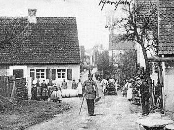Idylle in der Hauptstraße 1904, mit Gemeindeboten und Kuh-Gespann und einem Metzger mit Schürze, der das Schlachten vor dem Haus für die Aufnahme kurz unterbricht.