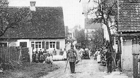 Idylle in der Hauptstraße 1904, mit Gemeindeboten und Kuh-Gespann und einem Metzger mit Schürze, der das Schlachten vor dem Haus für die Aufnahme kurz unterbricht.