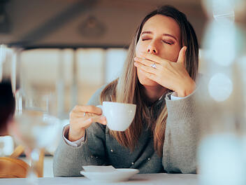 Gähnende Frau trinkt morgens Kaffee im Restaurant und wirkt müde Kaffee Gesundheit Müdigkeit Gähnen Restaurant Morgenroutine Koffein Abhängigkeit Toleranz Entzug Überkonsum Timing Nüchtern Magen Reflux Sodbrennen Zuckerzusatz Süßgetränk Energiecrash Schlafstörungen Nervosität Herzrasen Kopfschmerzen Dehydrierung Hydration Frühstück Achtsamkeit Pausen Stress Konzentration