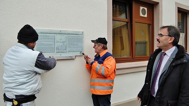 Die Bauhof-Mitarbeiter Thomas G&uuml;nter (links) und Werner Beck schrauben im Beisein von B&uuml;rgermeister Harald Hofmann (rechts) das neue Hinweisschild auf den Milit&auml;rhistorischen Wanderweg 1866 an die frisch verputzte Gasthauswand.  Foto: Sigismund von Dobsch&uuml;tz