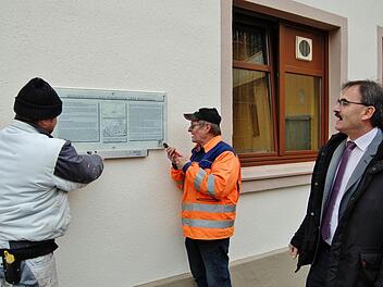 Die Bauhof-Mitarbeiter Thomas Günter (links) und Werner Beck schrauben im Beisein von Bürgermeister Harald Hofmann (rechts) das neue Hinweisschild auf den Militärhistorischen Wanderweg 1866 an die frisch verputzte Gasthauswand.  Foto: Sigismund von Dobschütz