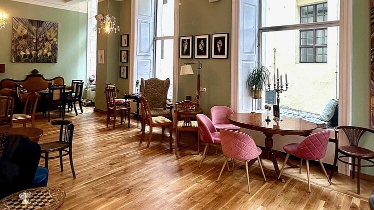 Helles Caf&eacute; mit Holzfu&szlig;boden und eleganter Einrichtung. Kronleuchter, gro&szlig;e Fenster, Kunstwerke an den W&auml;nden und verschiedene Sitzbereiche verleihen dem Raum eine gem&uuml;tliche Atmosph&auml;re.