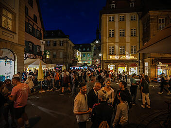 Menschenmengen füllen eine belebte, festlich beleuchtete Straße in einer Altstadt. Stände und Geschäfte, darunter "Käthe Wohlfahrt", sind zu sehen. Es herrscht eine fröhliche Atmosphäre.