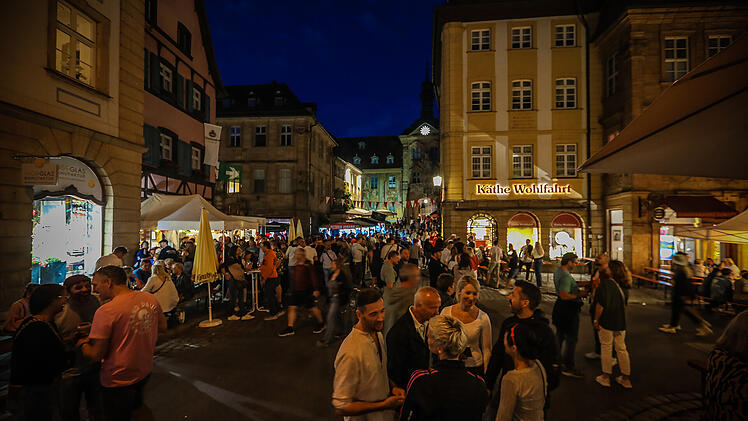 Menschenmengen füllen eine belebte, festlich beleuchtete Straße in einer Altstadt. Stände und Geschäfte, darunter "Käthe Wohlfahrt", sind zu sehen. Es herrscht eine fröhliche Atmosphäre.