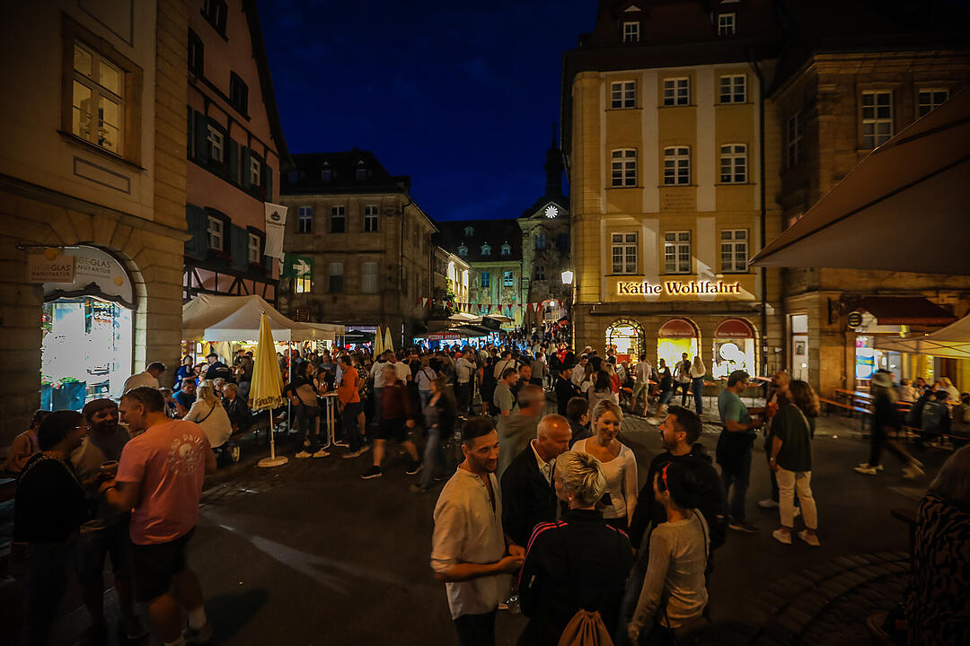 Menschenmengen füllen eine belebte, festlich beleuchtete Straße in einer Altstadt. Stände und Geschäfte, darunter "Käthe Wohlfahrt", sind zu sehen. Es herrscht eine fröhliche Atmosphäre.