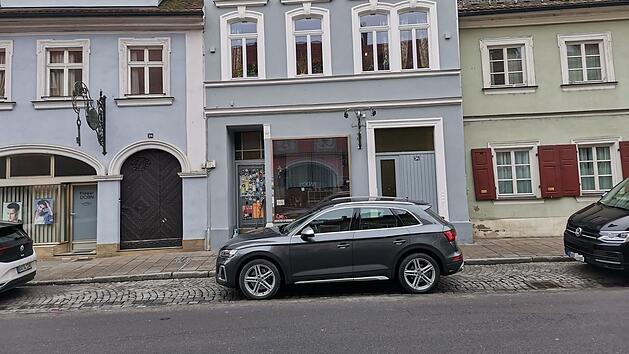 Eine graue Fassade mit gro&szlig;en Fenstern und einem Auto davor. Ein Schild &uuml;ber dem Eingang deutet auf die Bar hin. Umgebung hat klassische Architektur mit mehreren Schildern.