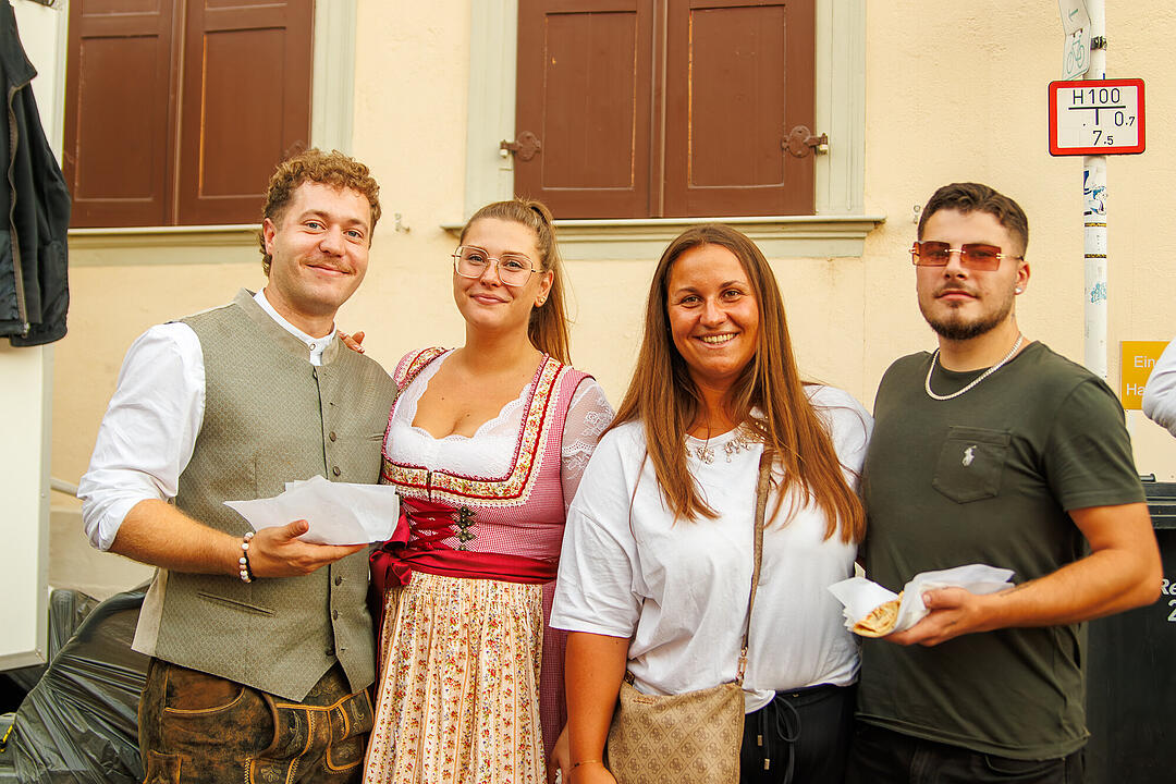 Vier Menschen genie&szlig;en die Sandkerwa in Bamberg. Eine Person tr&auml;gt Tracht, alle l&auml;cheln und halten Essen in den H&auml;nden. Hintergrund: eine Hauswand mit Fenstern.