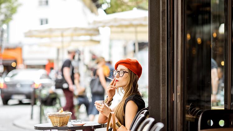 Junge stilvolle Frau mit roter Baskenmütze raucht eine Zigarette sitzen im Freien an der französischen Café Young stylish woman in red beret smoking a cigarette sitting outdoors at the french cafe