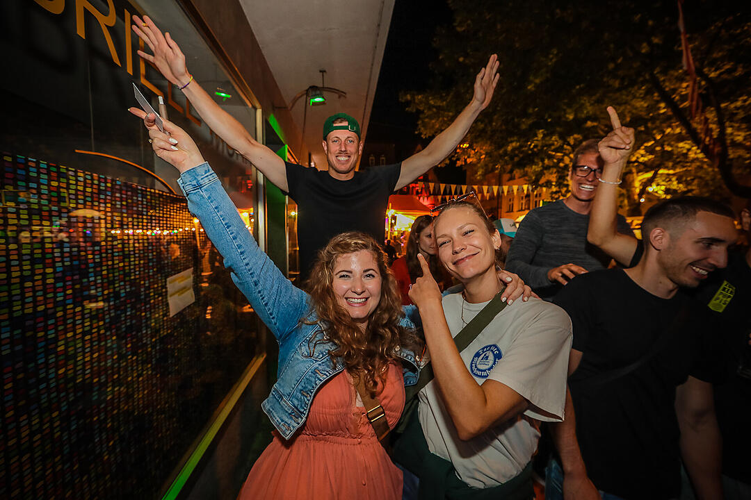 Menschen feiern fröhlich auf der Bamberger Sandkerwa 2025. Sie posieren lächelnd für ein Foto, von Festbeleuchtung und bunten Fahnen umgeben.