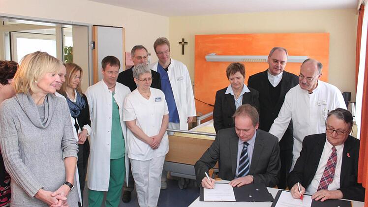 Landrat Eberhard Irlinger (vorne rechts) und Caritas-Geschäftsführer Markus Beck (daneben) unterzeichneten in einem Zimmer der Palliativstation des Höchstadter Krankenhauses den Kooperationsvertrag. Im Hintergrund alle, die auf dieser Station aktiv sind.