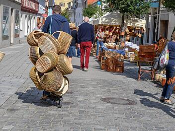 Eine belebte Straße auf dem Lichtenfelser Korbmarkt zeigt Stände mit Körben. Menschen schlendern vorbei, während ein Mann viele Körbe transportiert.