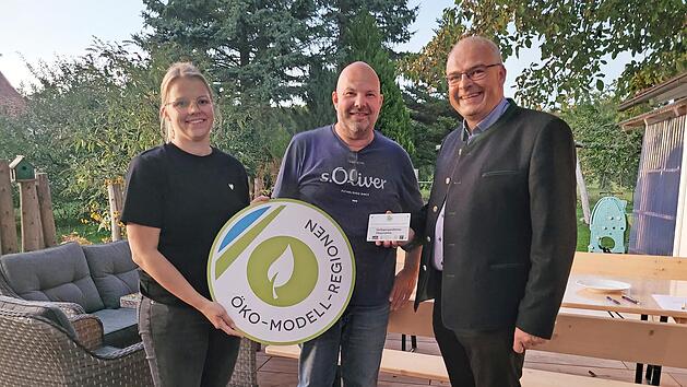 Die Öko-Modellregionmanagerin Laura Pircher und Gößweinsteins Bürgermeister Hanngörg Zimmermann (r.) übergeben die Förderplakette an Stefan Simmerlein.