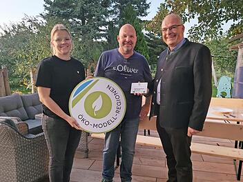 Die Öko-Modellregionmanagerin Laura Pircher und Gößweinsteins Bürgermeister Hanngörg Zimmermann (r.) übergeben die Förderplakette an Stefan Simmerlein.