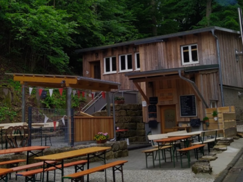 Ein traditioneller Biergarten mit Holztischen und -bänken im Freien, umgeben von Wald. Ein Gebäude aus Holz im Hintergrund. Bunte Wimpel hängen über dem Bereich.