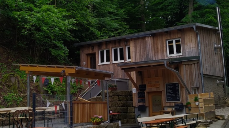 Ein traditioneller Biergarten mit Holztischen und -bänken im Freien, umgeben von Wald. Ein Gebäude aus Holz im Hintergrund. Bunte Wimpel hängen über dem Bereich.