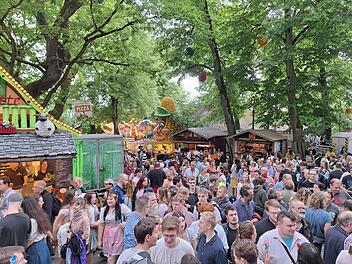 Belebtes Fest mit vielen Menschen im Grünen, umgeben von Buden und Ständen. Ein festlicher Anlass mit sommerlicher Atmosphäre und lebendiger Szenerie in einem bewaldeten Bereich.