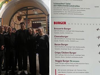 Eine Gruppe steht vor einem Restaurant. Rechts ist ein Men&uuml; mit durchgestrichenen Preisen, darunter handgeschriebene, niedrigere Preise mit Edding.