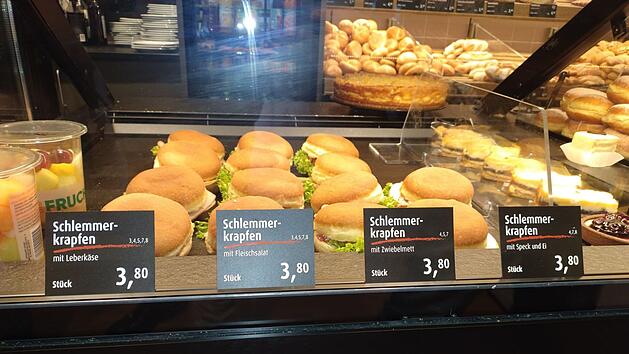 Ein B&auml;ckereidisplay zeigt Schlemmerkrapfen mit Leberk&auml;se, Fleischsalat, Zwiebelmett, Speck und Ei, alle zu 3,80 &euro;. Links sind Obstbecher zu sehen.