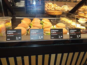 Ein Bäckereidisplay zeigt Schlemmerkrapfen mit Leberkäse, Fleischsalat, Zwiebelmett, Speck und Ei, alle zu 3,80 €. Links sind Obstbecher zu sehen.
