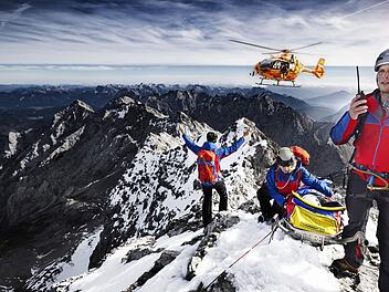 Auch die zweite Staffel der "Bergretter im Einsatz" zeigt Bergwachteins&auml;tze in den bayerischen Alpen - hautnah und in beeindruckenden Bildern. Drei Angeh&ouml;rige der Bergwacht, die sich mit einer geretteten Person auf einem Berggipfel befinden, weisen einen anfliegenden Hubschrauber ein.