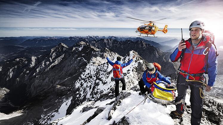 Auch die zweite Staffel der "Bergretter im Einsatz" zeigt Bergwachteins&auml;tze in den bayerischen Alpen - hautnah und in beeindruckenden Bildern. Drei Angeh&ouml;rige der Bergwacht, die sich mit einer geretteten Person auf einem Berggipfel befinden, weisen einen anfliegenden Hubschrauber ein.