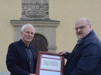 Kirchenpfleger Heinz Kraus (links) und Rainer Domke zeigen die Originalurkunde von 1357 im Holzrahmen. Im Hintergrund das Westportal der Simultankirche Theisenort mit der bisher falsch interpretierten Jahreszahl. Foto: eh