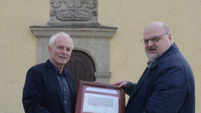 Kirchenpfleger Heinz Kraus (links) und Rainer Domke zeigen die Originalurkunde von 1357 im Holzrahmen. Im Hintergrund das Westportal der Simultankirche Theisenort mit der bisher falsch interpretierten Jahreszahl. Foto: eh